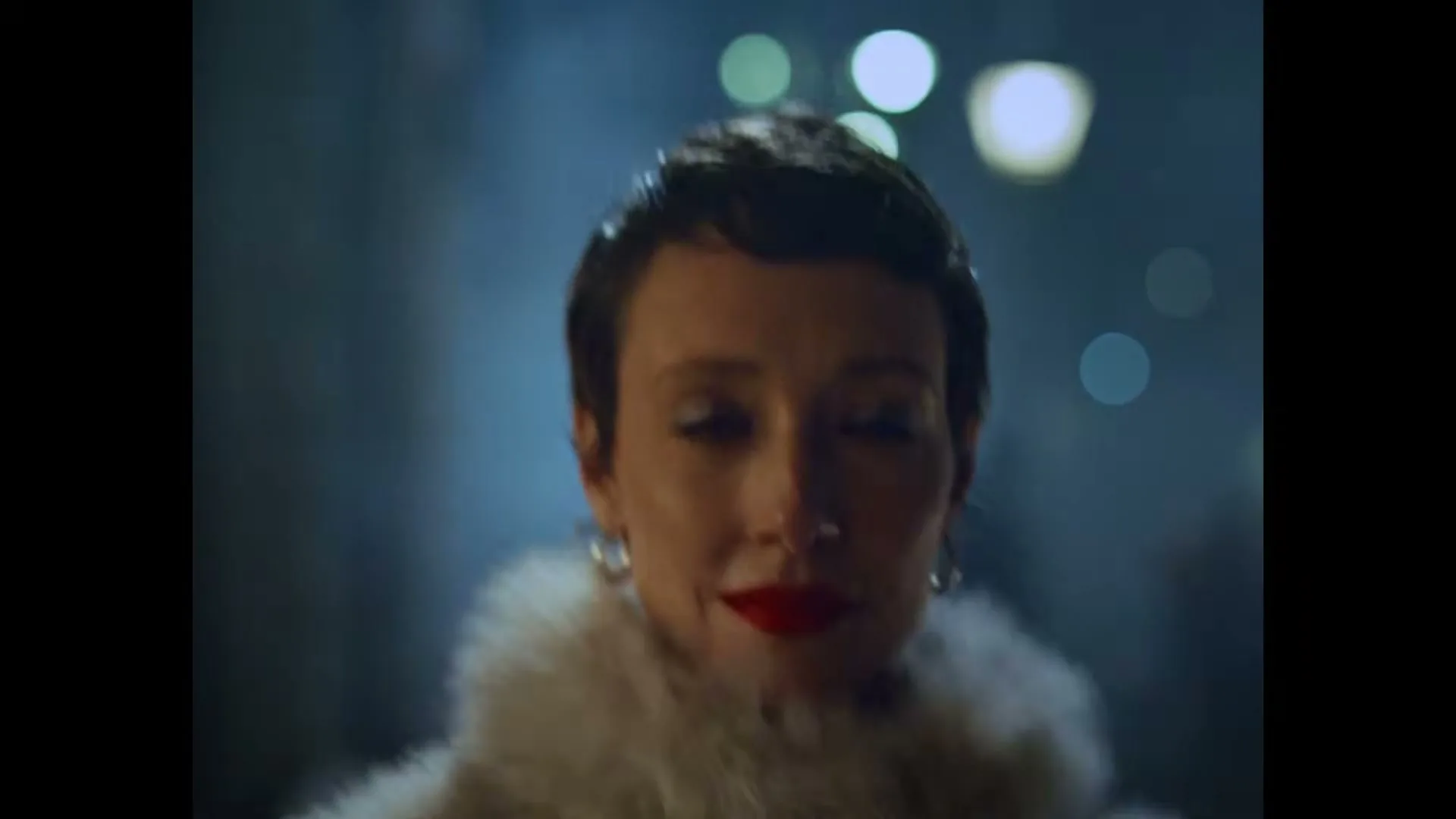A woman with short dark hair and red lipstick, wearing a white fur coat, looks up and smiles in a blurry, dark urban setting at night.