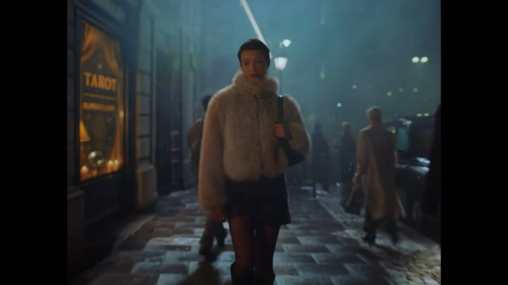 A woman in a white fur coat and black skirt walks confidently down a dark, misty city street at night, passing a tarot shop.