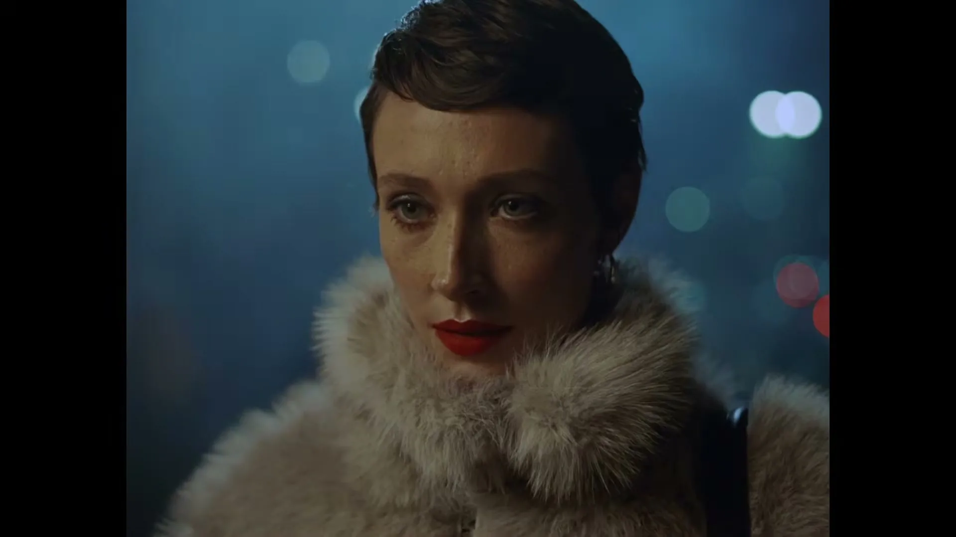 A woman with short dark hair and red lipstick, wearing a fur coat, looks up with a neutral expression at night.