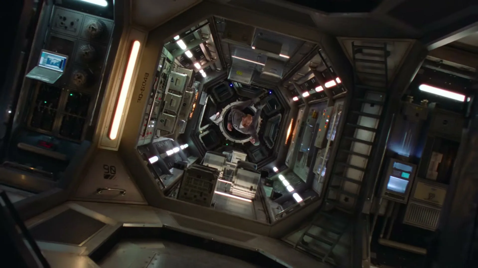 A man in a grey jumpsuit floats in zero gravity inside a futuristic, hexagonal spaceship corridor, reaching out as if to grab something.