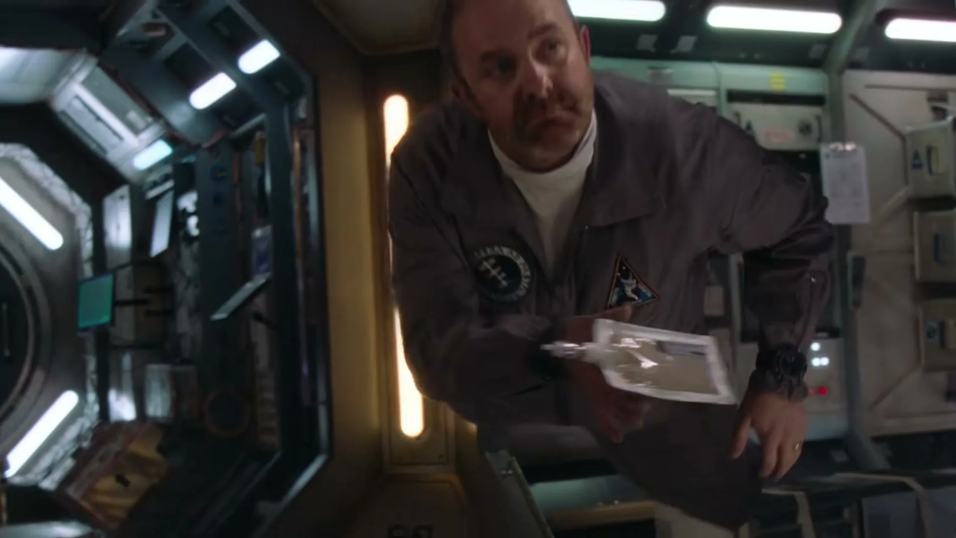 A male astronaut in a grey jumpsuit floats in a spaceship corridor, holding a food pouch, looking around with a curious expression.
