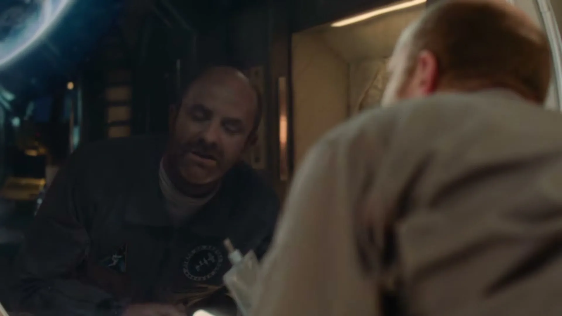 A bald man in a dark uniform holds a pouch and talks to another person, reflected in a round window of a spaceship.