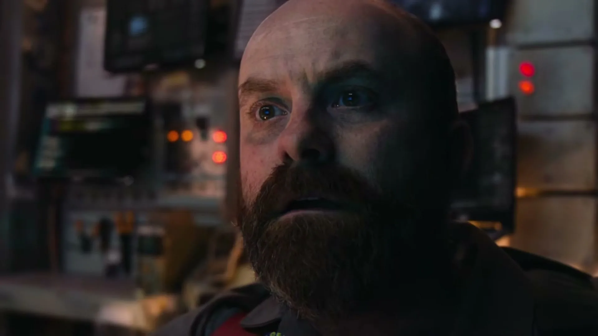 A bald man with a beard looks up with a worried expression in a dimly lit control room, surrounded by screens and buttons.