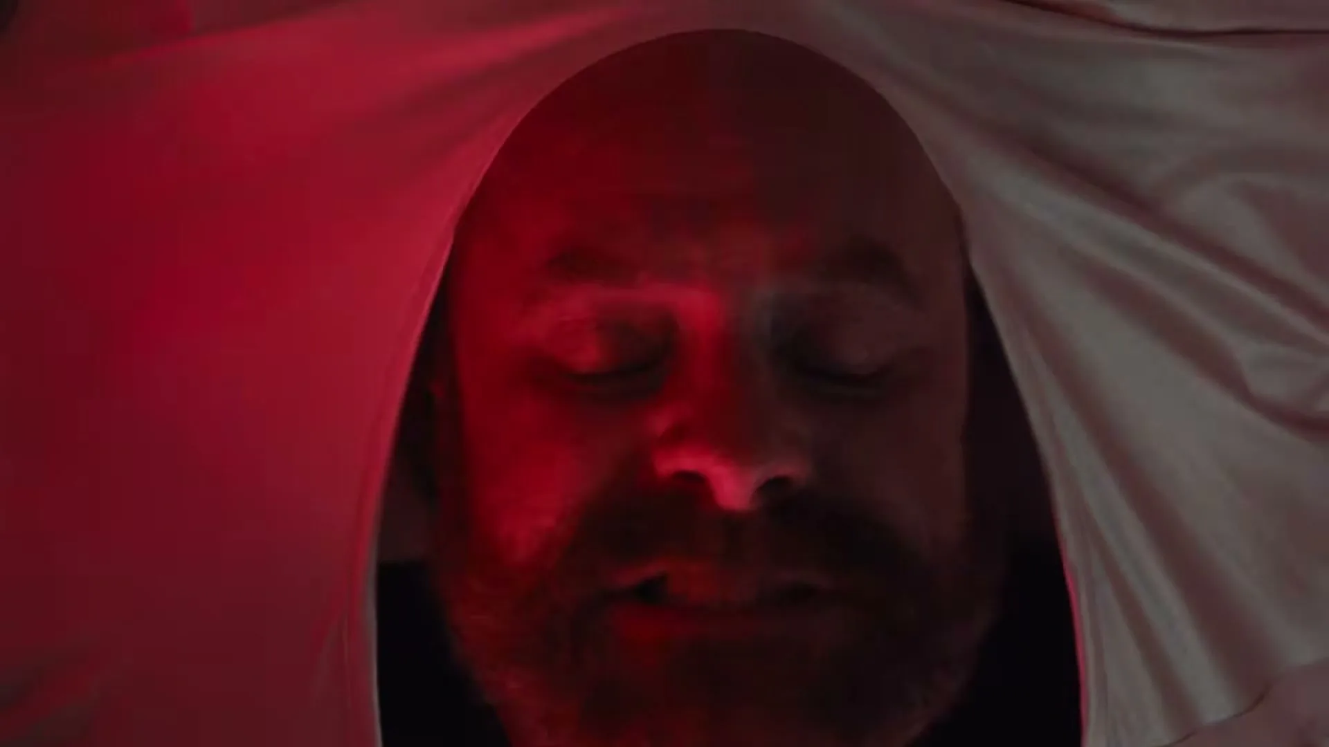 A bearded man with closed eyes and an open mouth is seen in a closeup shot, illuminated by a red light under a white sheet.