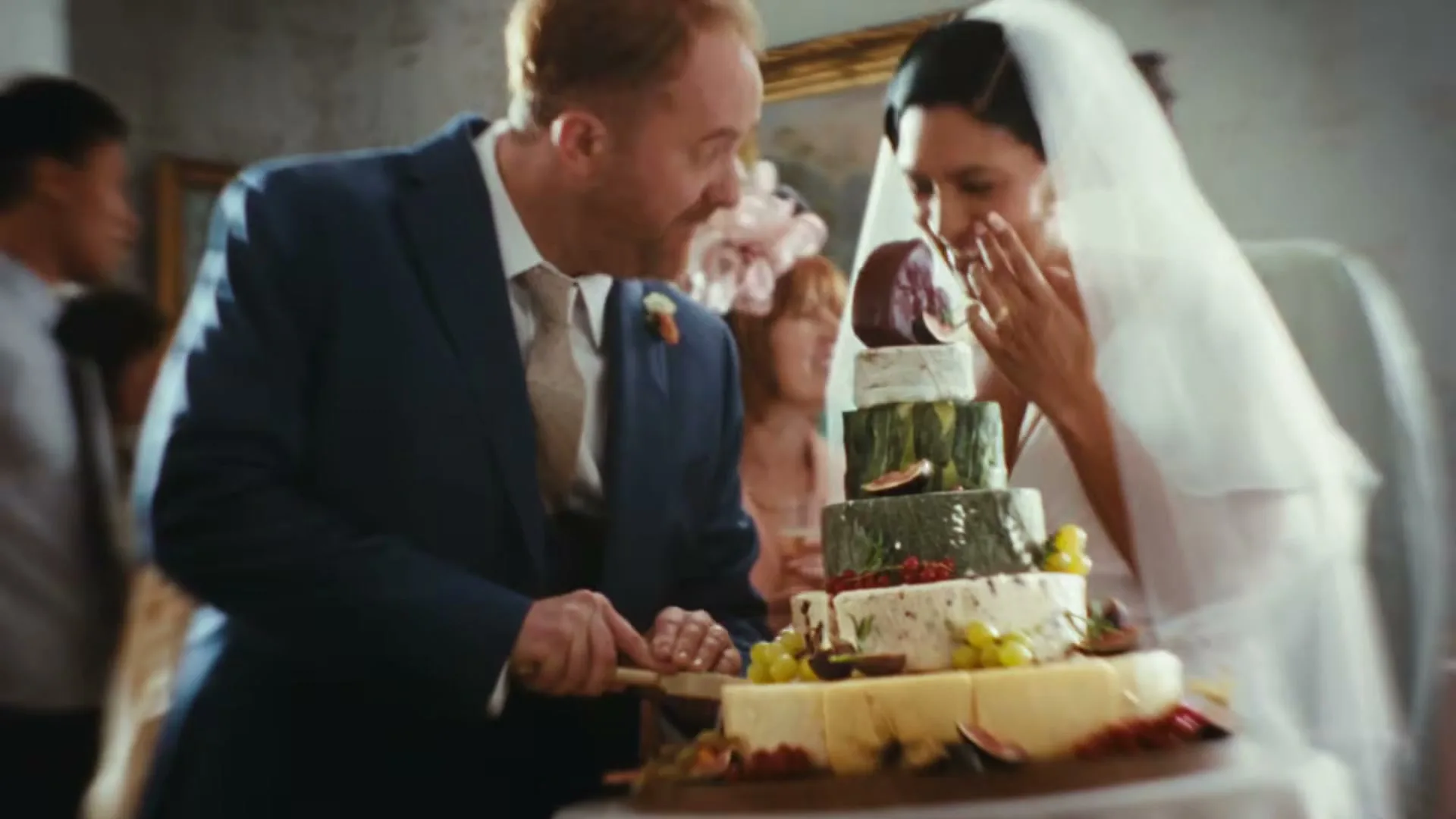 A ginger-haired groom in a blue suit and a bride in a white veil cut a tiered cheese wedding cake, smiling at each other in a grand interior.