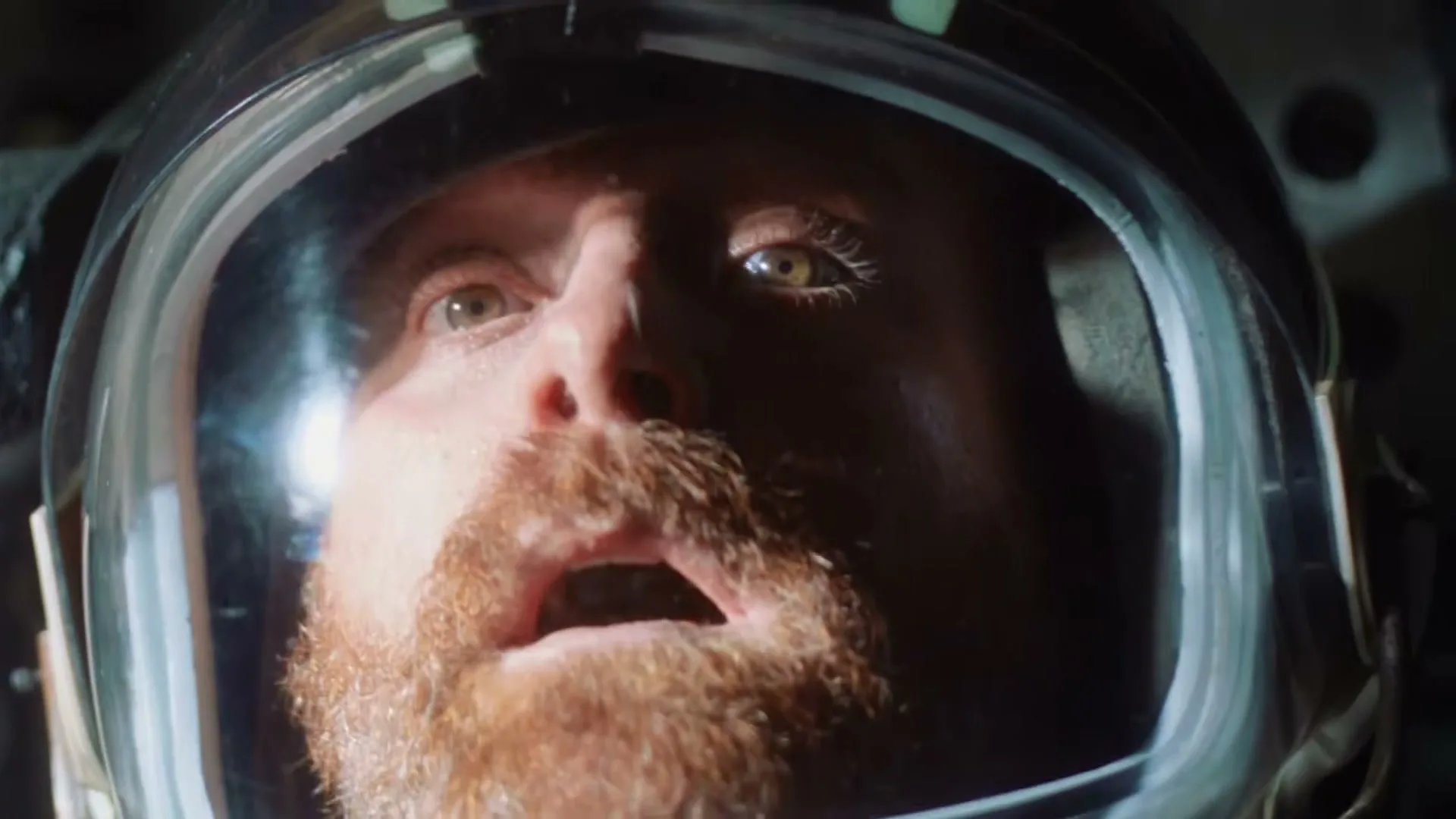A male astronaut with a red beard opens his eyes in surprise while wearing a helmet in a spaceship.