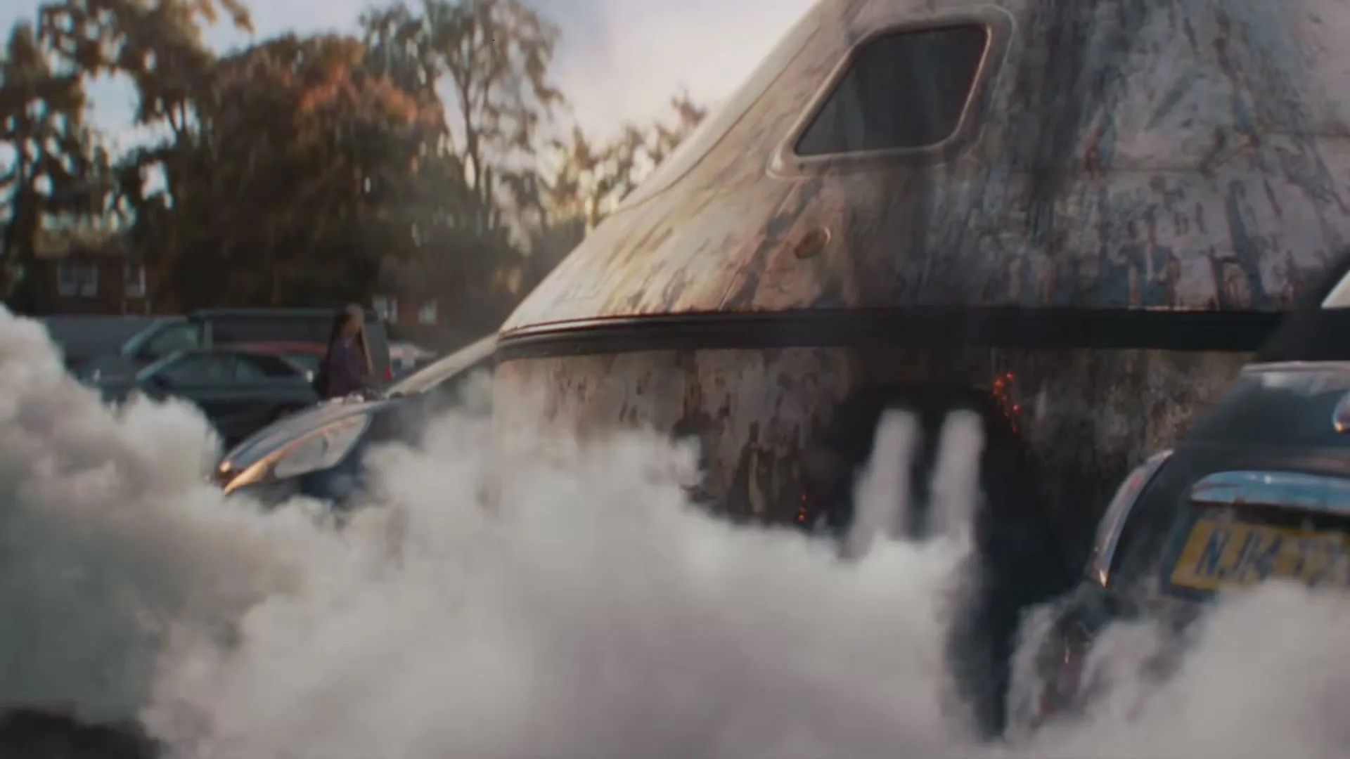 A large, burnt, metallic space capsule lands in a parking lot, emitting smoke and steam, surrounded by cars and a distant person.