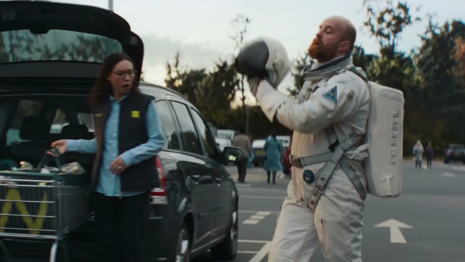 A man in an astronaut suit takes off his helmet next to a car in a parking lot, while two women look at him.