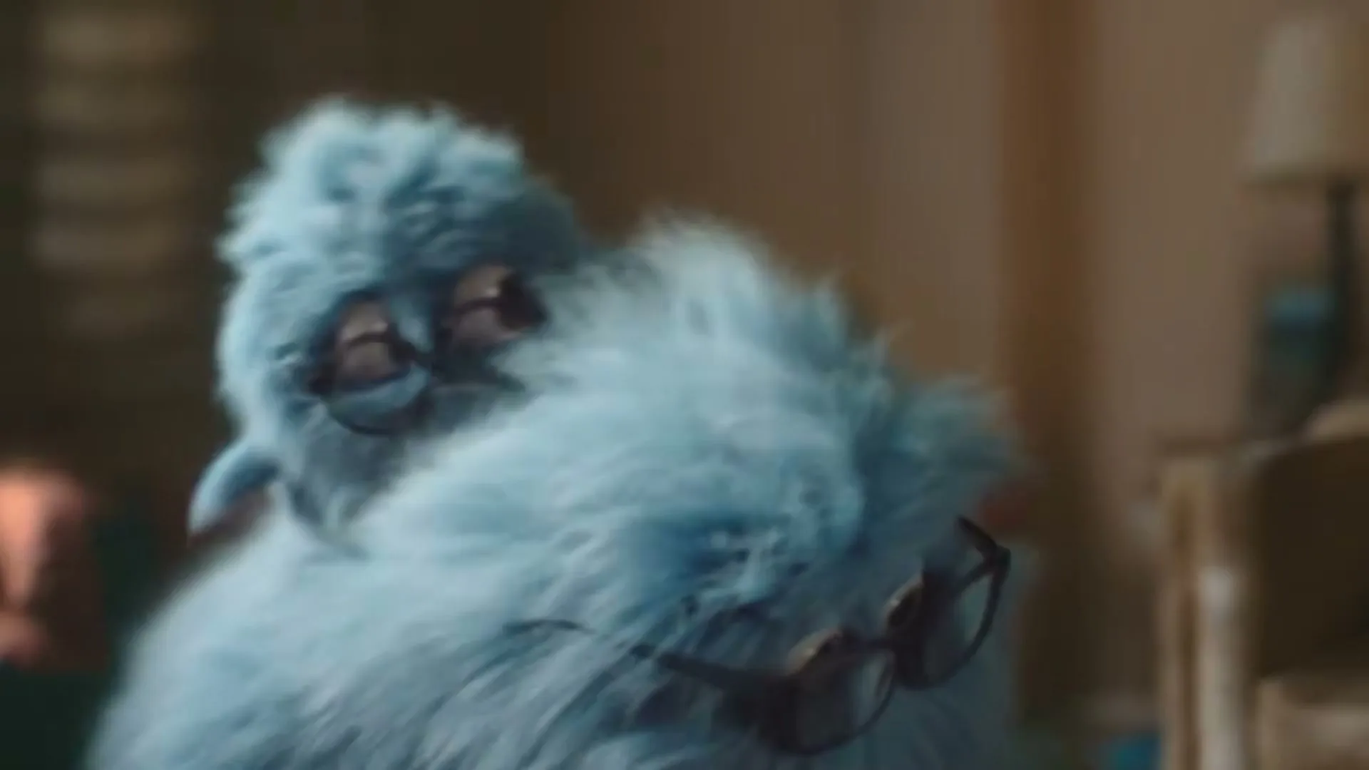 A fluffy blue monster with glasses on its head and chest looks around a dimly lit room.