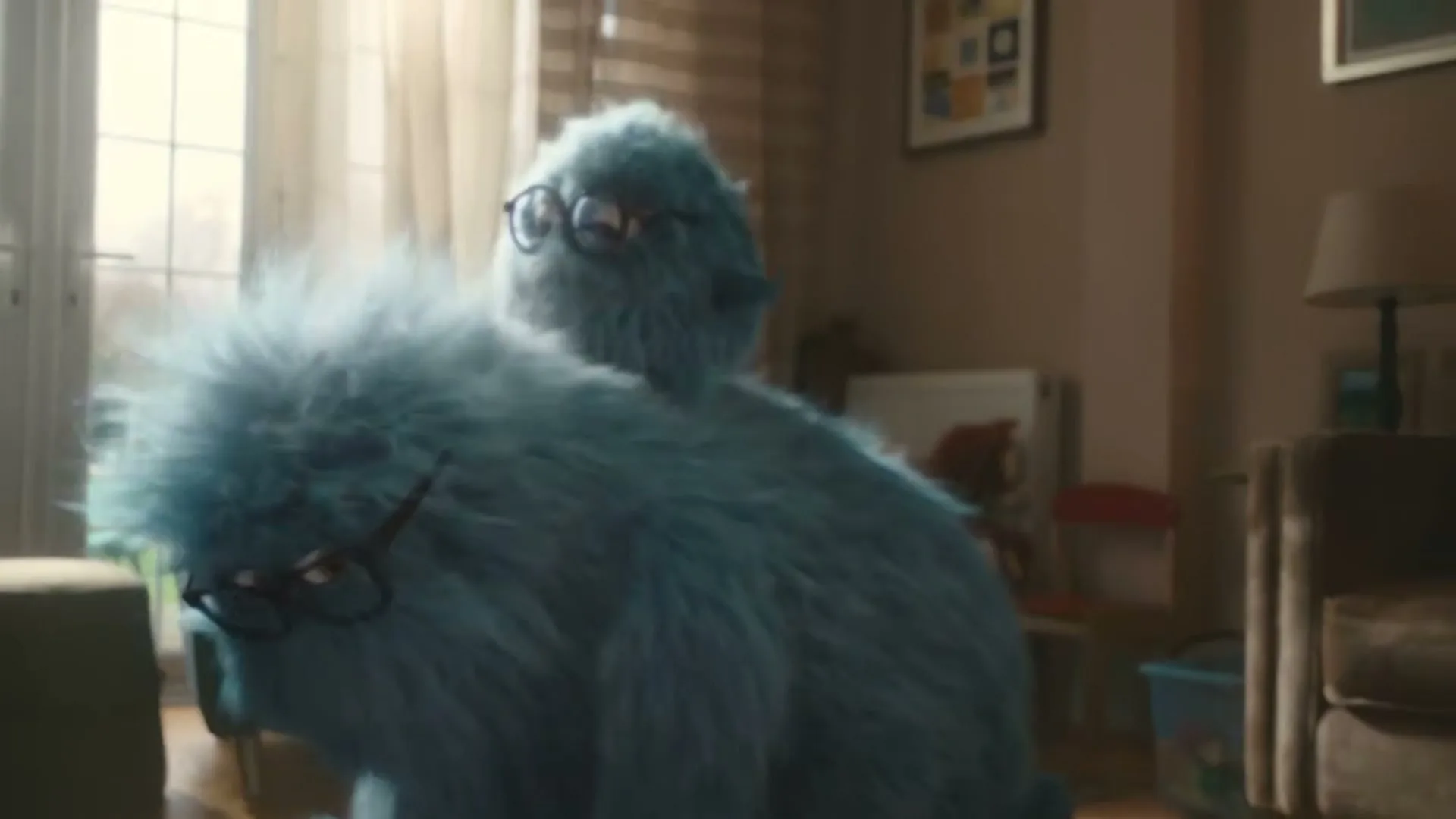 Two blue, furry monsters, one large and one small, with glasses, are in a living room, with the smaller monster riding on the back of the larger one.