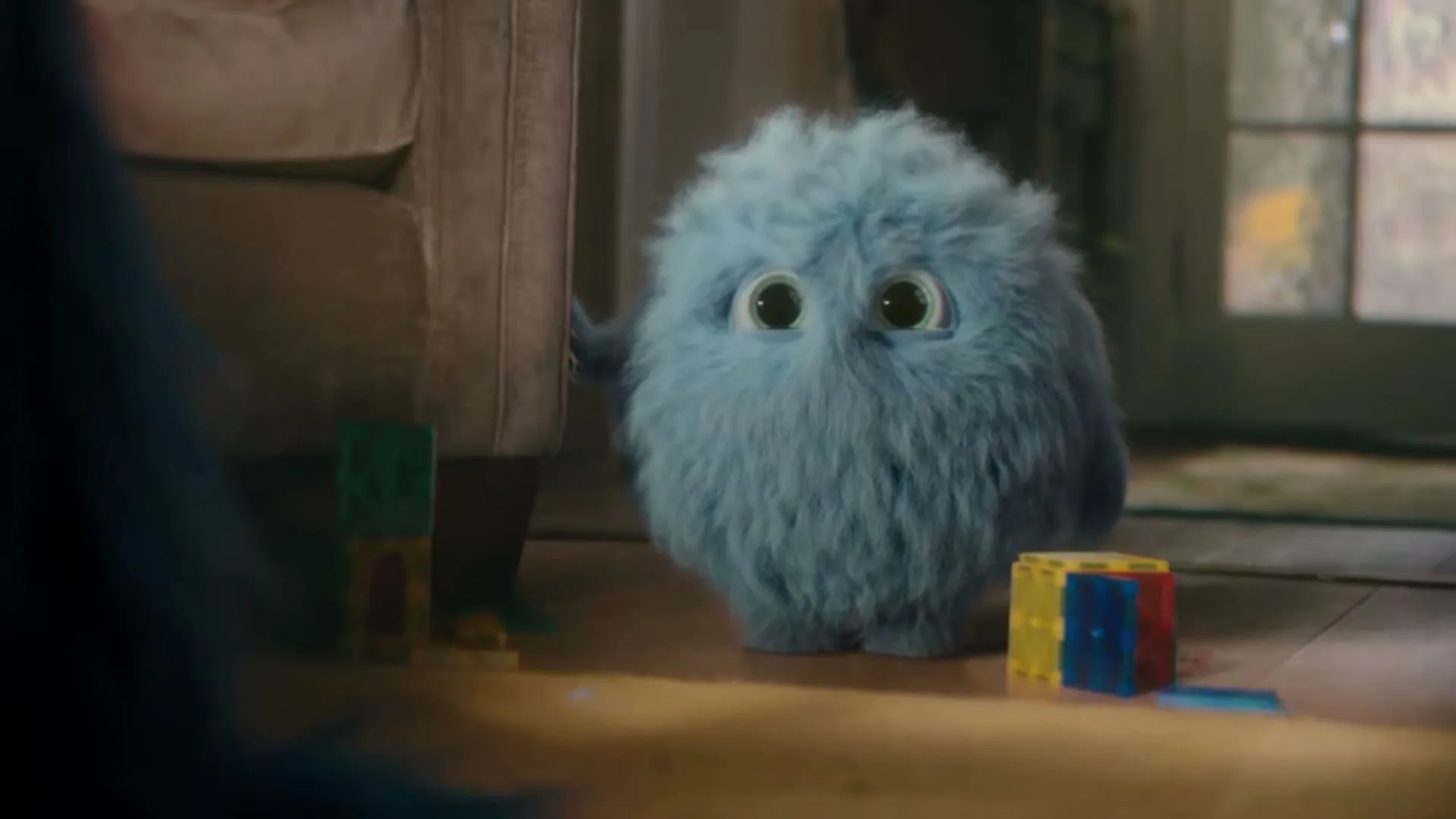A small, fluffy blue creature with big eyes peeks from behind a sofa in a living room, looking curious.