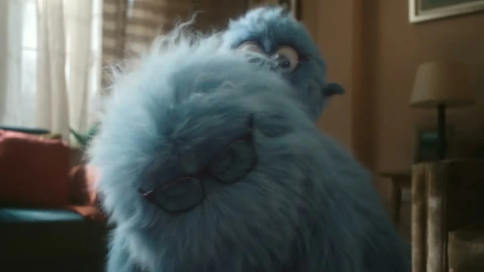 A large, fluffy blue monster with glasses on its head looks around a living room, with a smaller blue monster peeking over its shoulder.