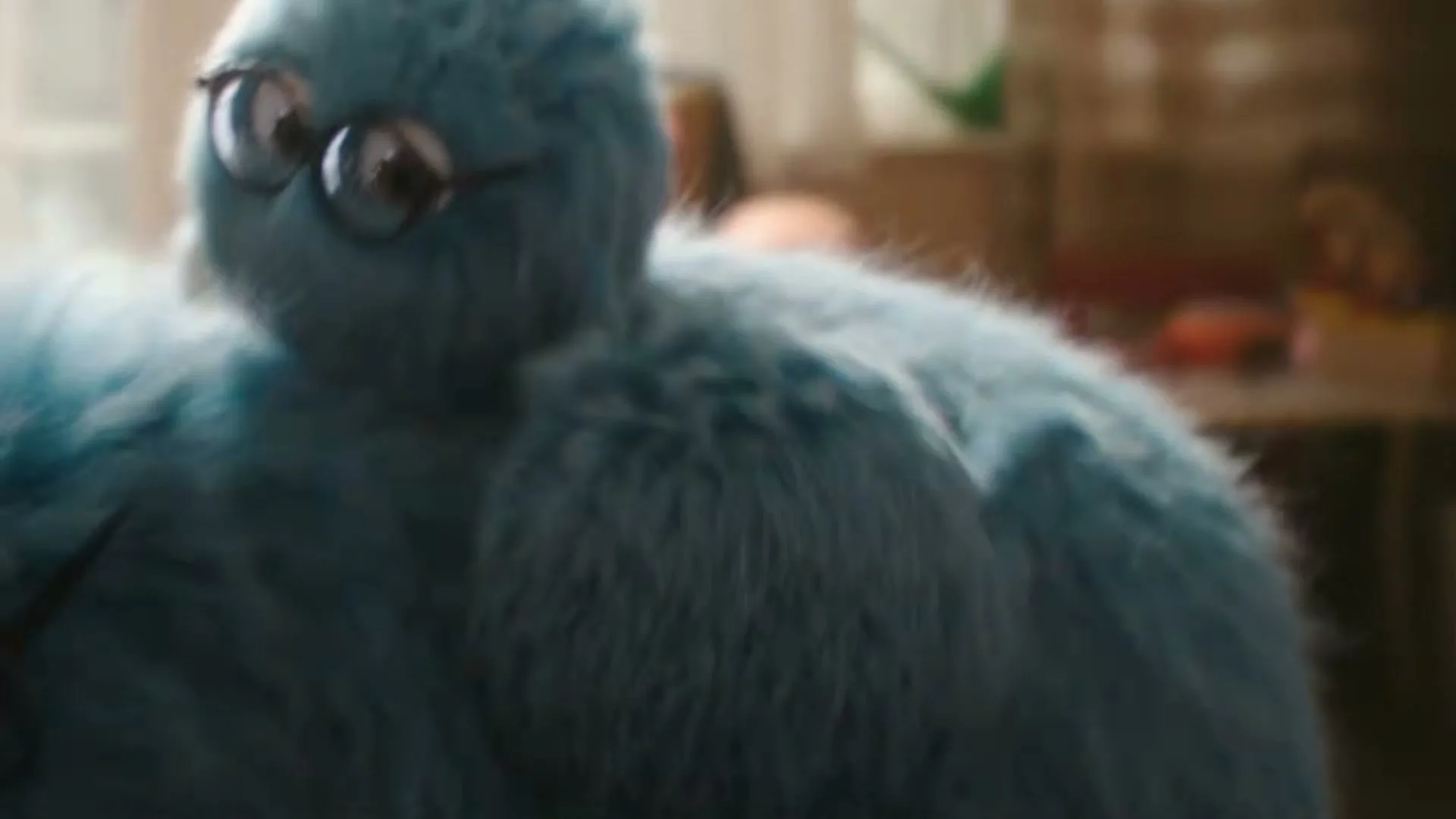 A small, round, fluffy blue creature with glasses walks towards a larger, similar creature, then snuggles into its side in a cozy interior.