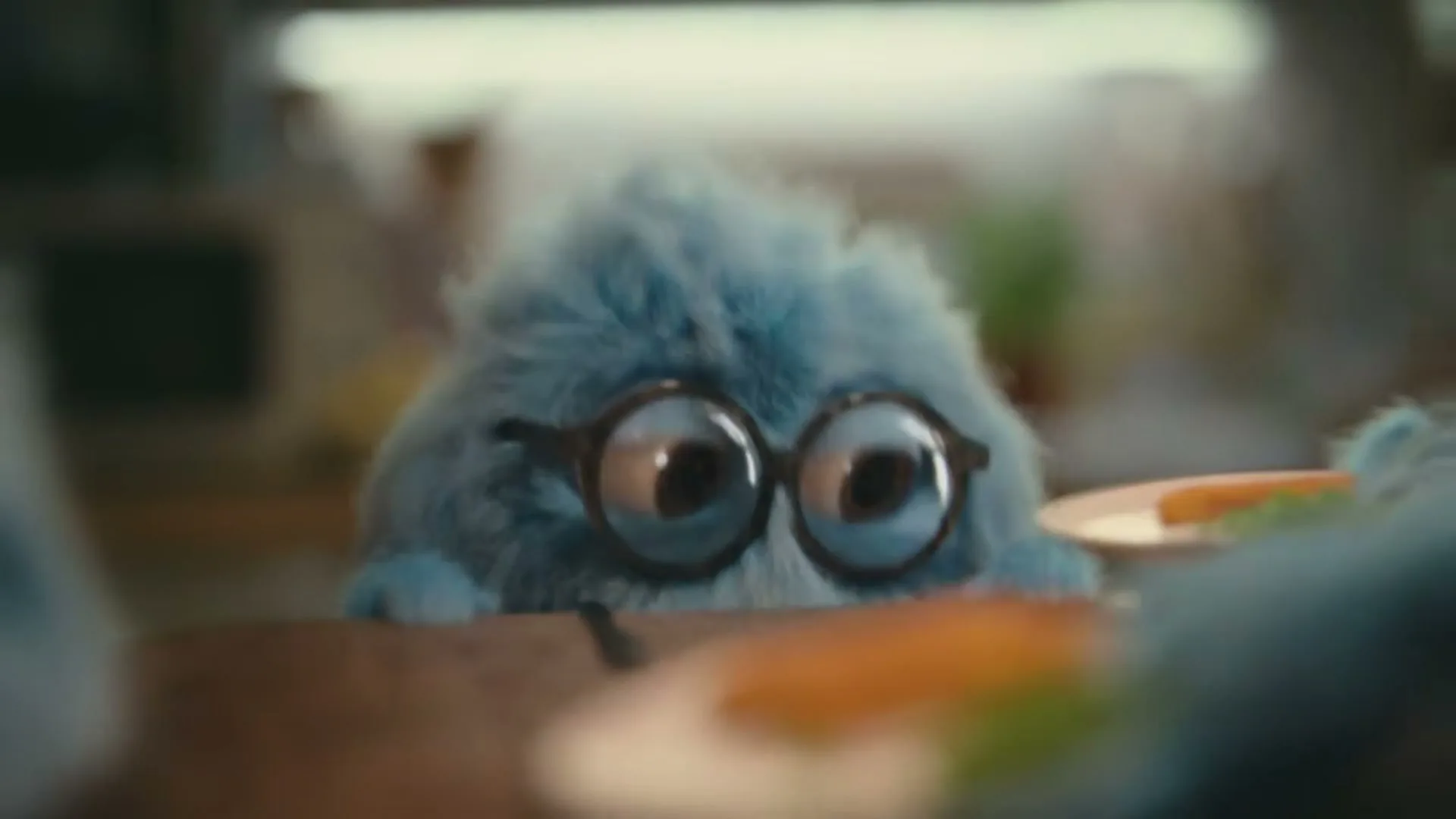 A small, fluffy blue creature with large glasses peeks over a wooden table in a blurry kitchen setting.