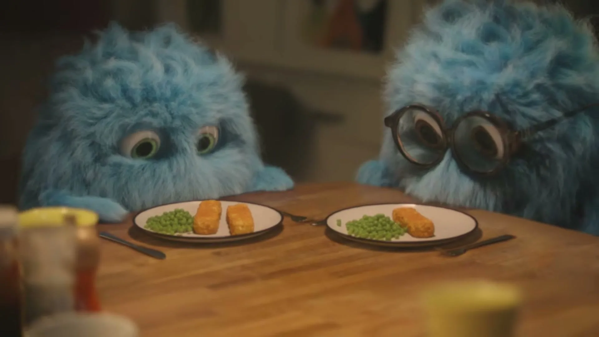Two blue furry creatures, one with glasses, sit at a wooden table looking at plates of fish sticks and peas.
