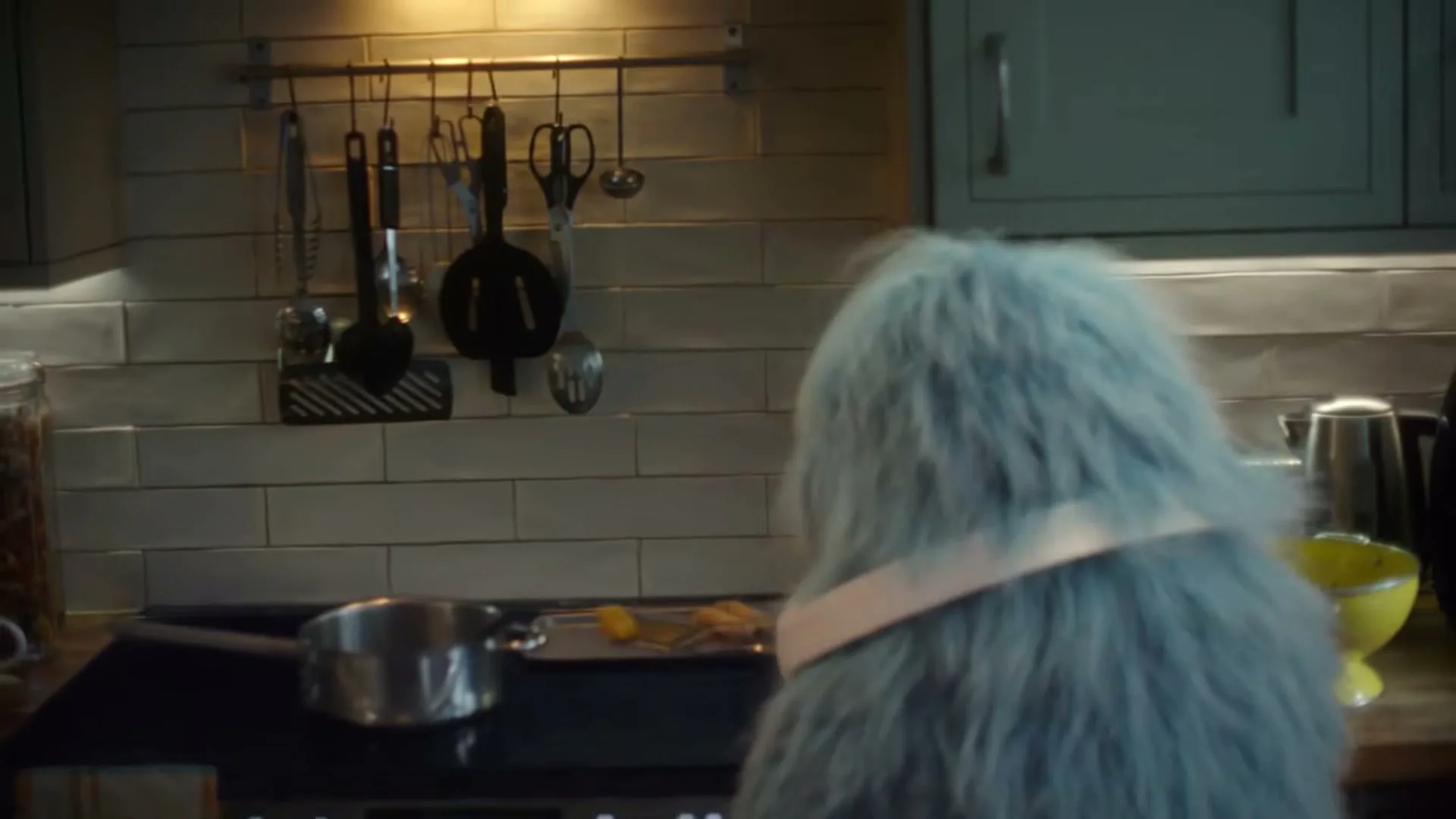 A furry blue creature with a pink collar stands at a stove in a kitchen, flipping food with a spatula.