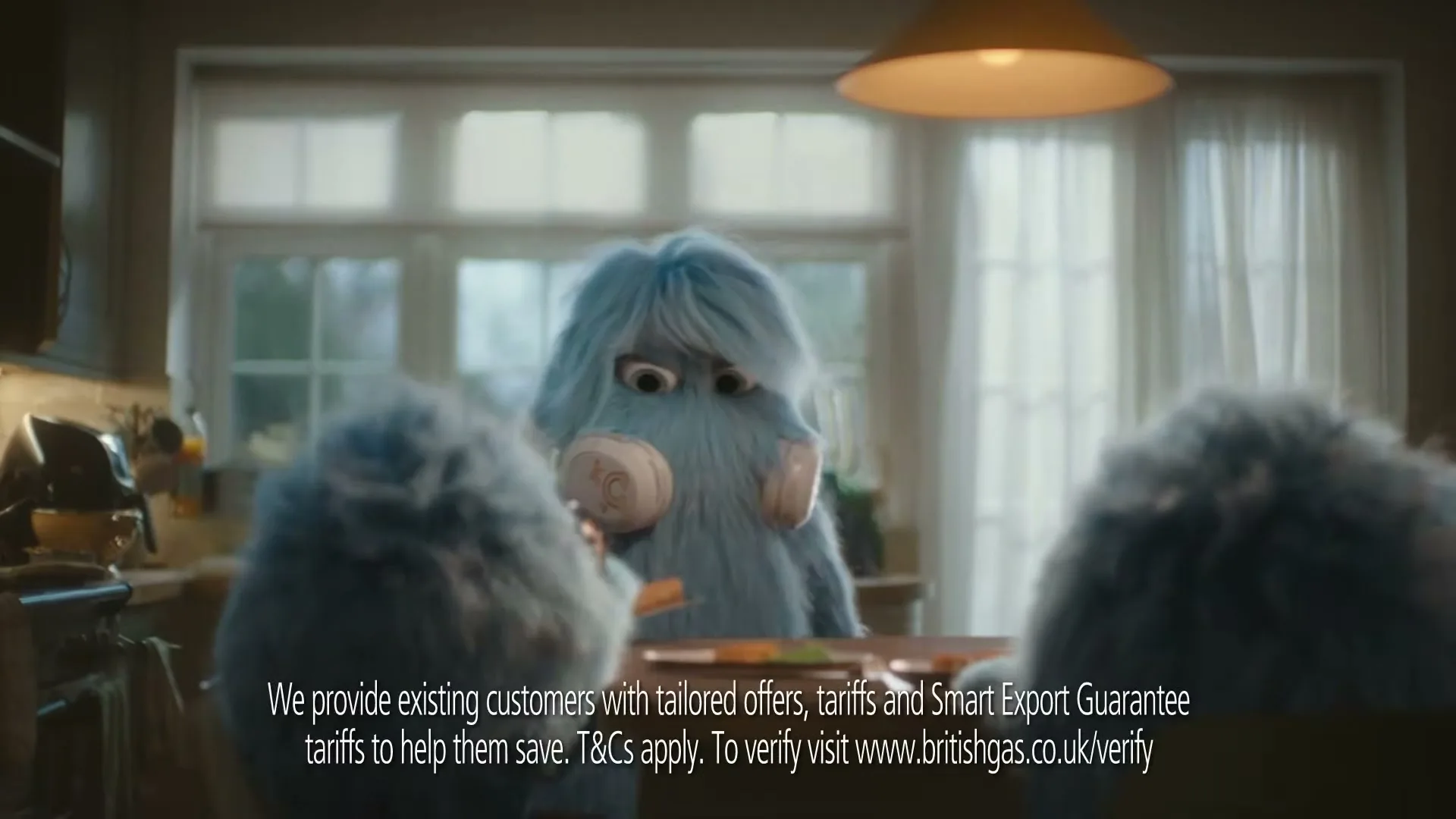 A blue furry creature with headphones sits at a kitchen table, looking down at something, with two other furry creatures partially visible in the foreground.