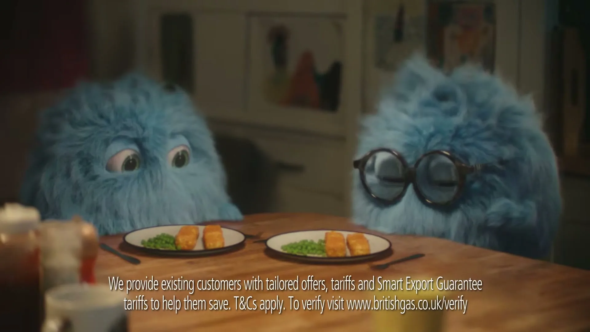 Two blue, furry, animated creatures with big eyes sit at a wooden table, each with a plate of fish sticks and peas, in a cozy home.