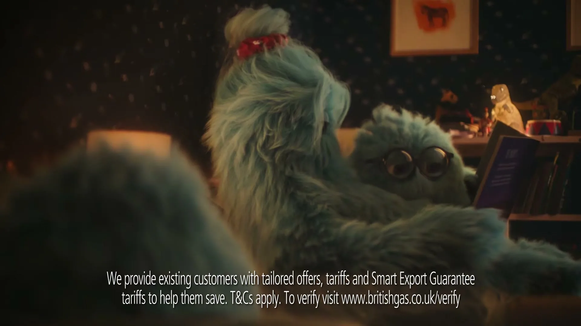Two fluffy blue monsters, one with glasses, are snuggled together on a sofa, reading a book in a cozy living room at night.
