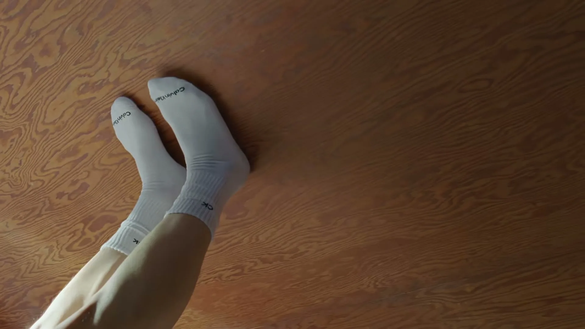 A person's legs and feet, wearing white Calvin Klein socks, are seen against a wooden floor, with the camera looking down.