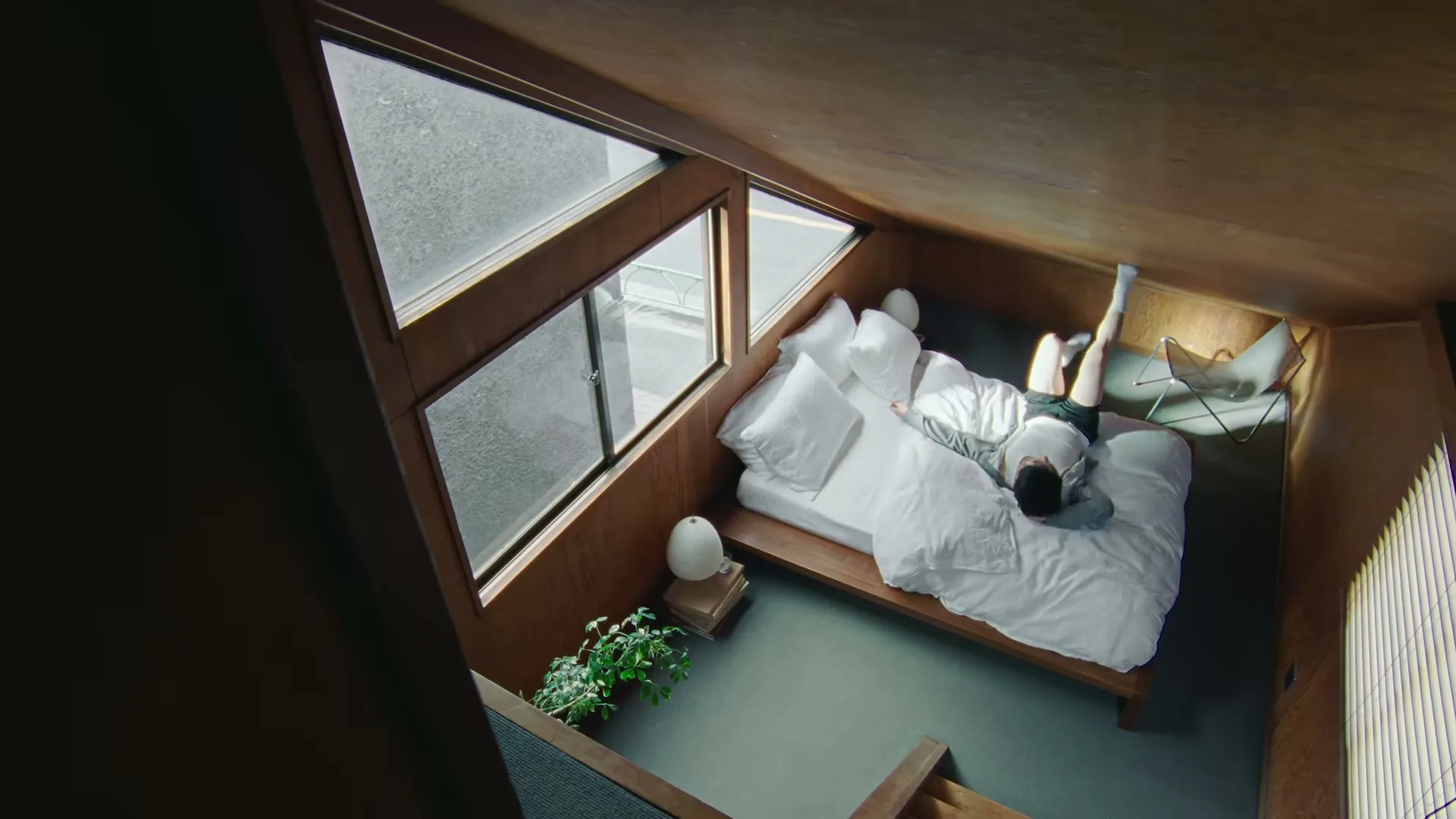 A young man in a grey hoodie and white socks lies on a bed in a modern, wood-paneled bedroom with large windows, then sits up.