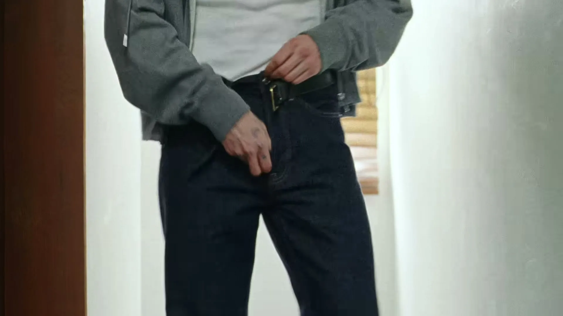 A man in a grey hoodie and dark jeans fastens his belt in a brightly lit room with a window.