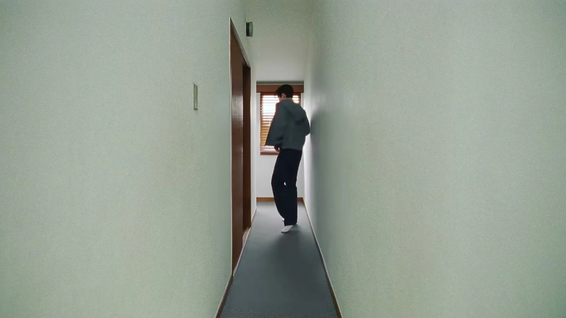 A young man in a grey hoodie and dark pants walks down a narrow hallway, taking off his hoodie.