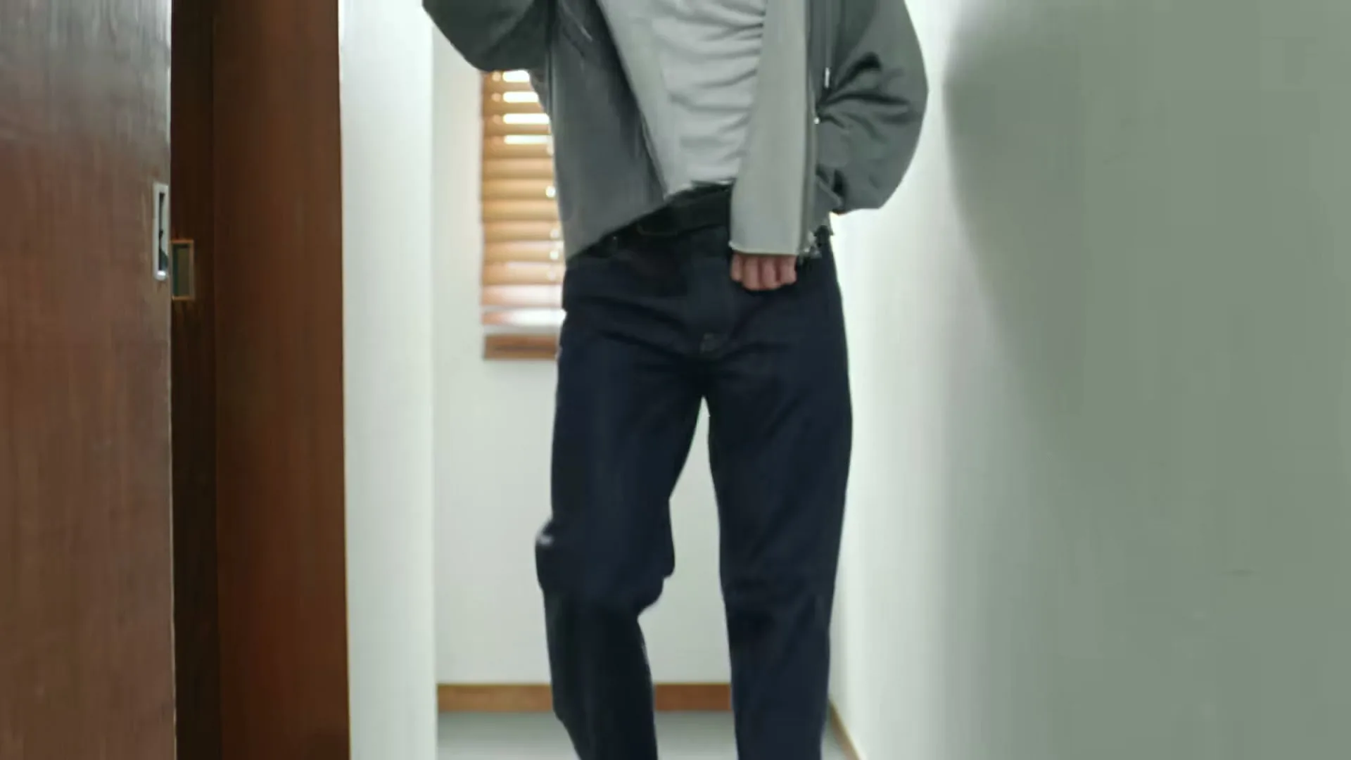A person in a grey hoodie and dark jeans walks down a hallway, adjusting their clothing, in a modern interior setting.