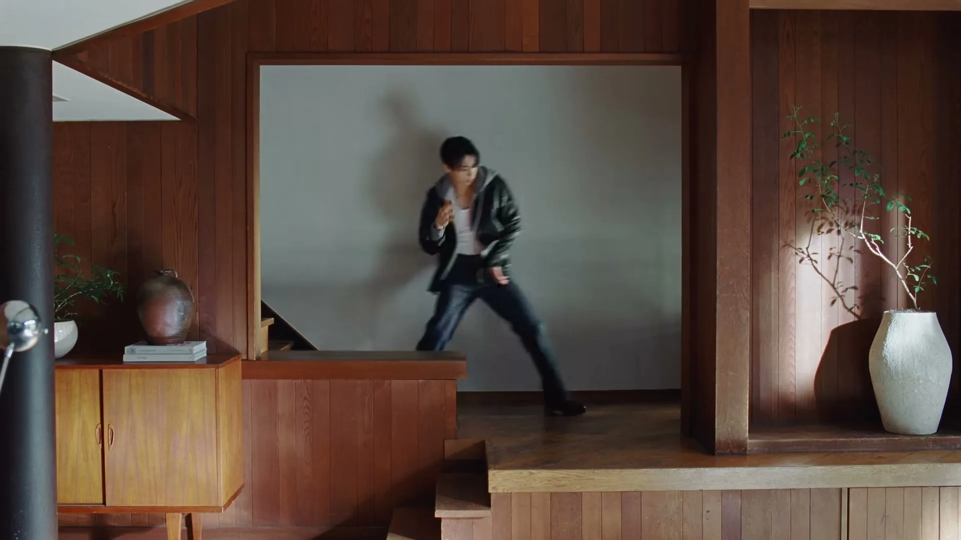 A young man in a leather jacket and jeans dances energetically in a modern, wood-paneled interior with white walls and plants.
