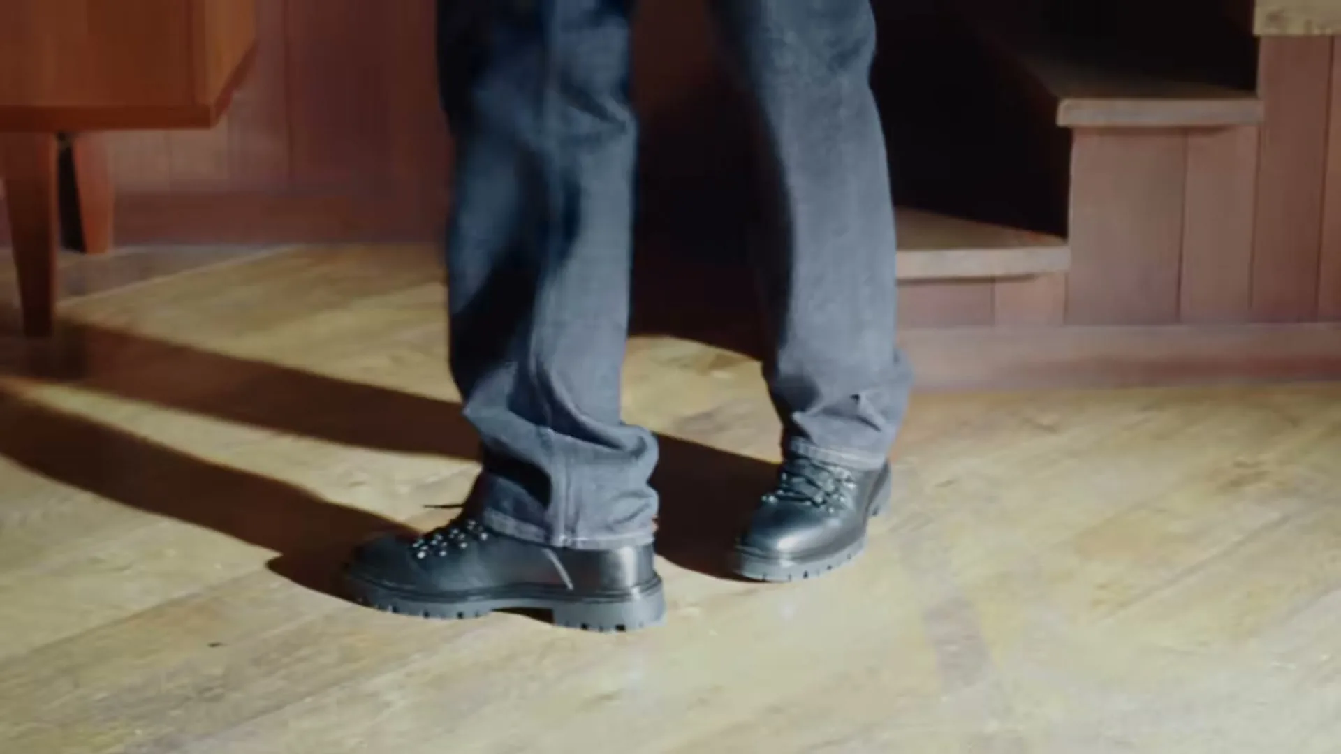 A person wearing dark jeans and black boots walks on a wooden floor, with a wooden staircase in the background.