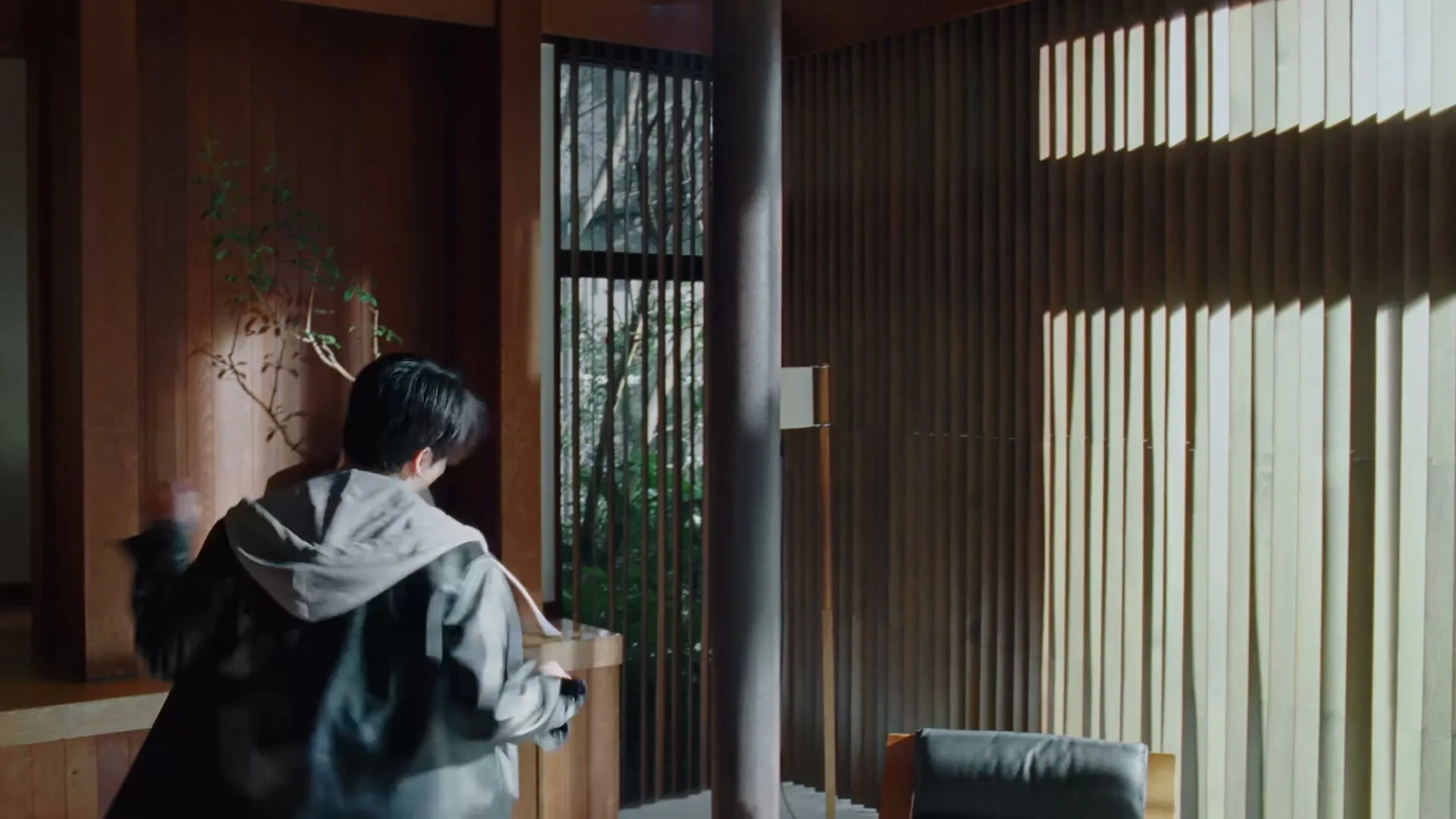 A young man in a grey hoodie and black jacket walks quickly through a modern, wood-paneled room with sunlight streaming through a window.
