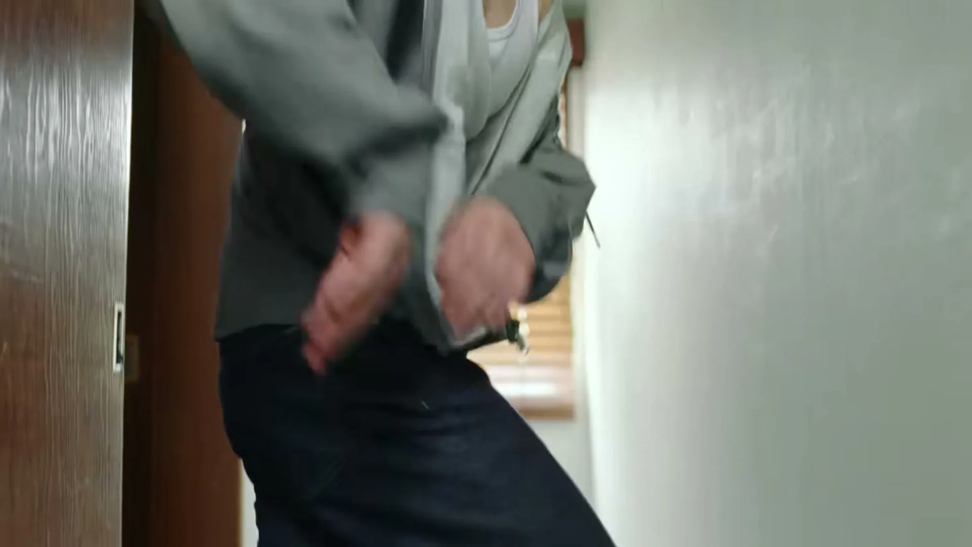 A person in a grey jacket and dark jeans is seen from the waist down, quickly moving their hands to zip up their jacket in an interior hallway.