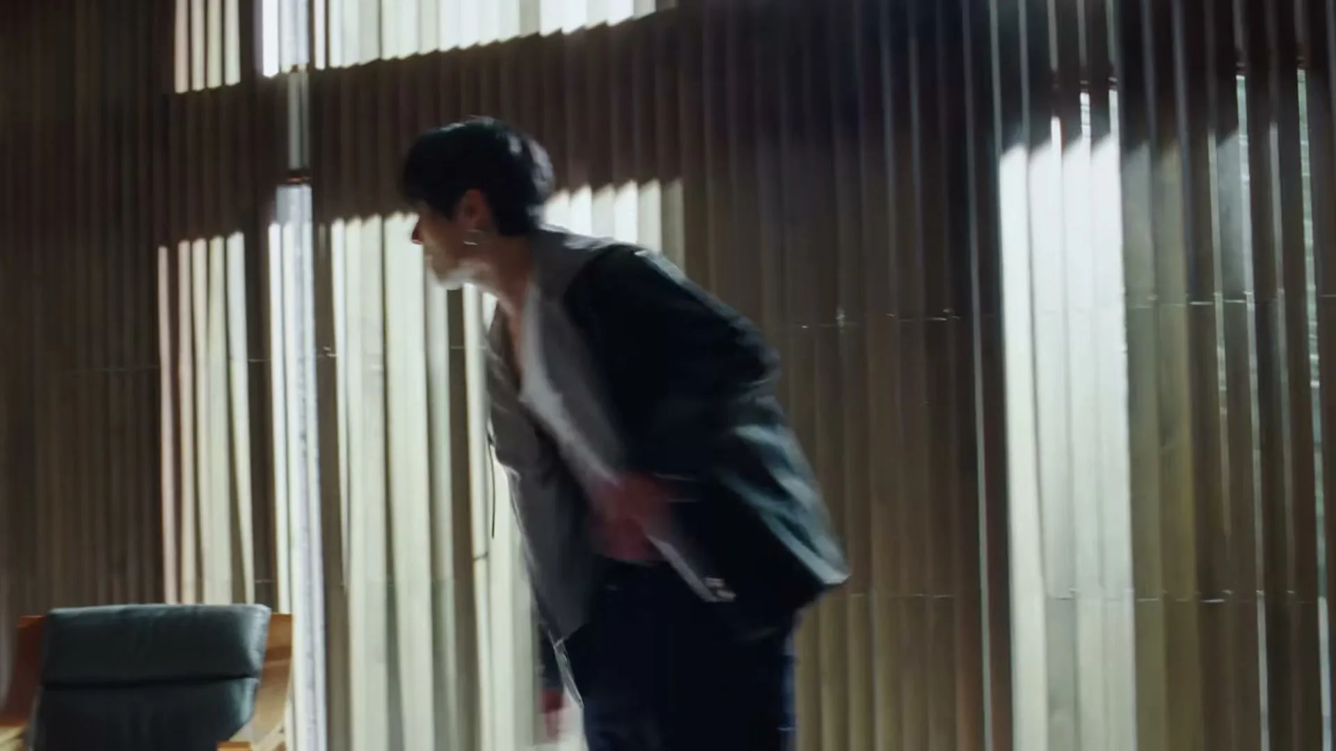 A young man with dark hair, wearing a black jacket and jeans, walks quickly across a sunlit room with vertical blinds.