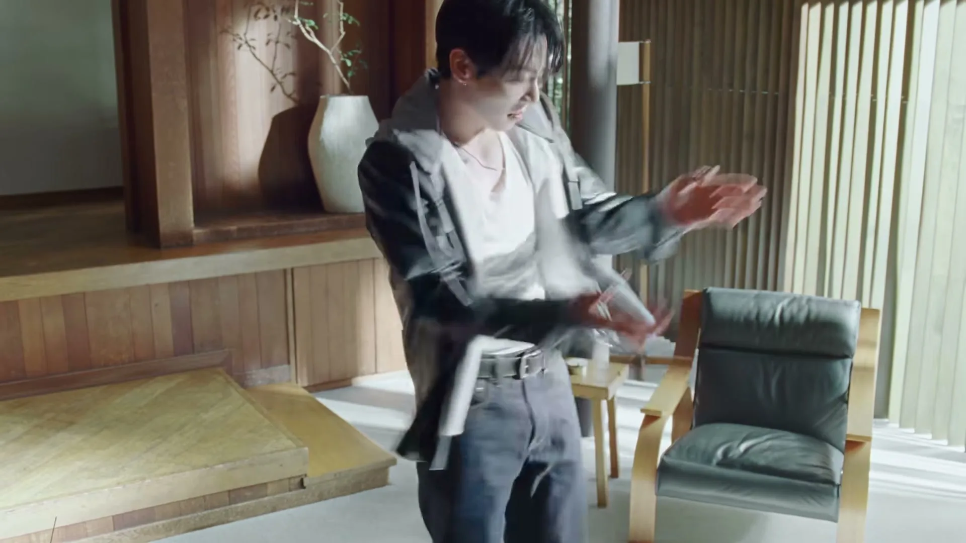 A young man in a white tank top and dark jeans puts on a black leather jacket in a modern, wood-paneled room with a chair and plant.