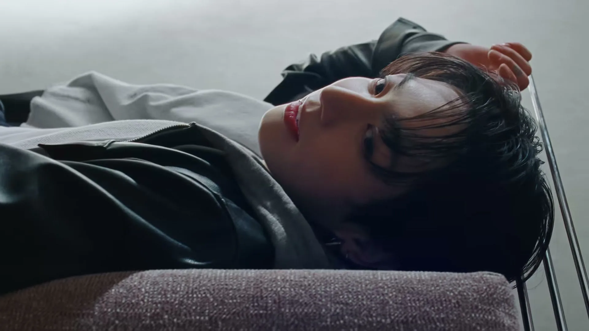 A young man with dark hair and a black leather jacket lies on a couch, looking up with a relaxed expression in an interior setting.