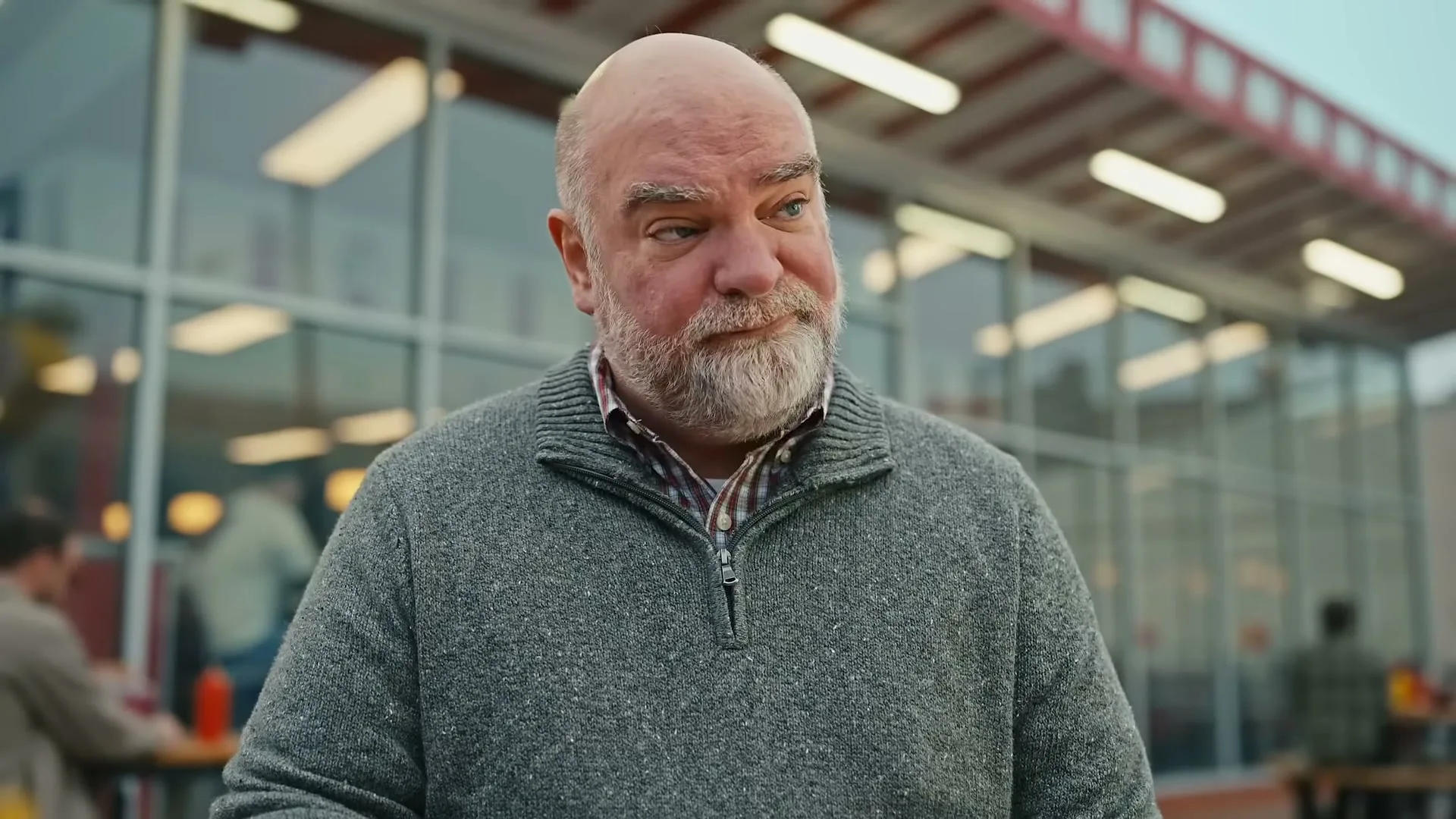 A bald, bearded man in a grey sweater looks at a burger on a tray, then up with a thoughtful expression, at an outdoor restaurant.