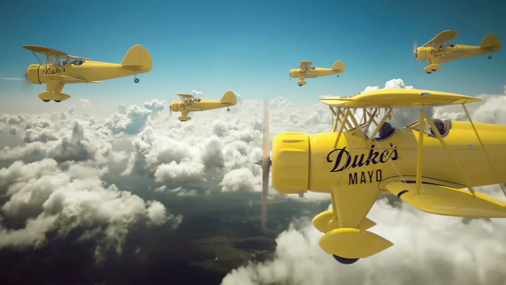 A squadron of five yellow biplanes, each with "Duke's Mayo" on its side, flies through a bright blue sky above white clouds.