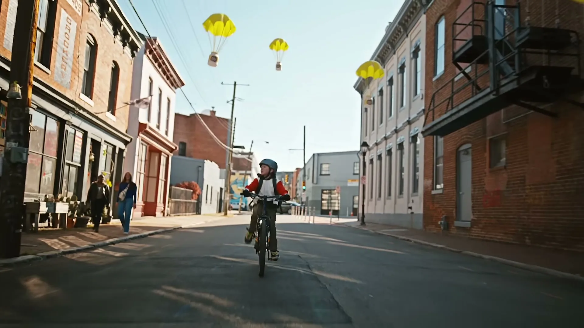 A young boy in a helmet and red jacket rides a bicycle down a city street, looking up at several yellow parachutes carrying packages descending from the sky.