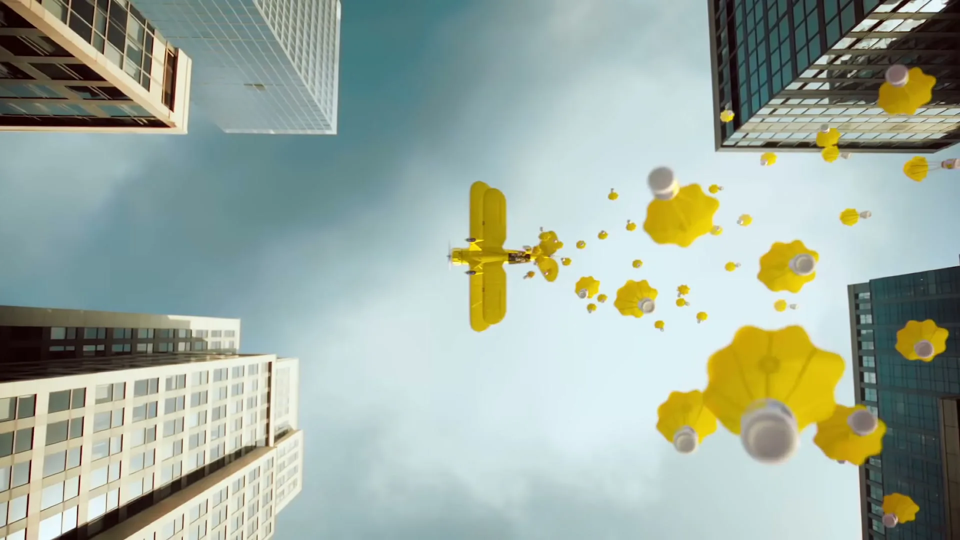 A yellow biplane flies through a city skyline, dropping yellow and white parachutes, seen from a low angle looking up at the sky.