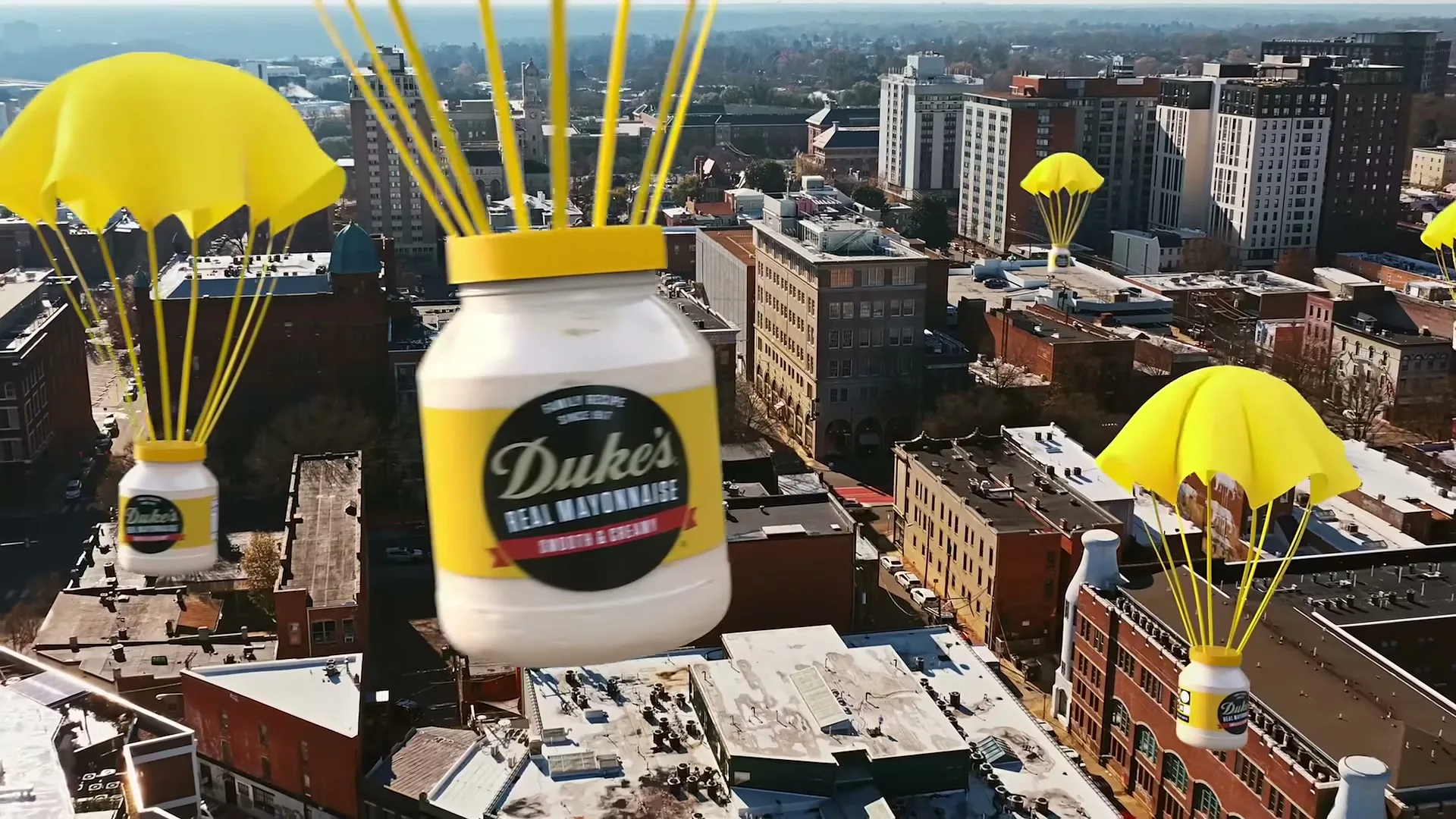 Dukes mayonnaise jars with yellow parachutes descend over a city skyline with buildings and rooftops under a clear sky.
