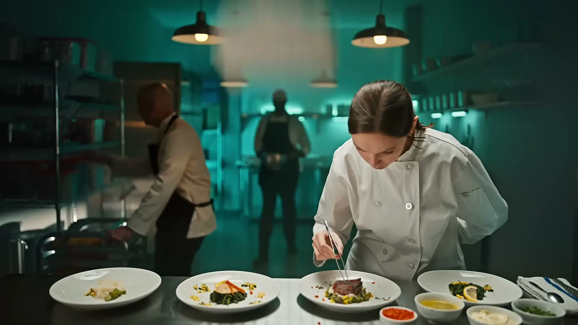 A female chef in a white uniform meticulously garnishes plates in a restaurant kitchen, then looks up in surprise as a jar of mayonnaise explodes above her.