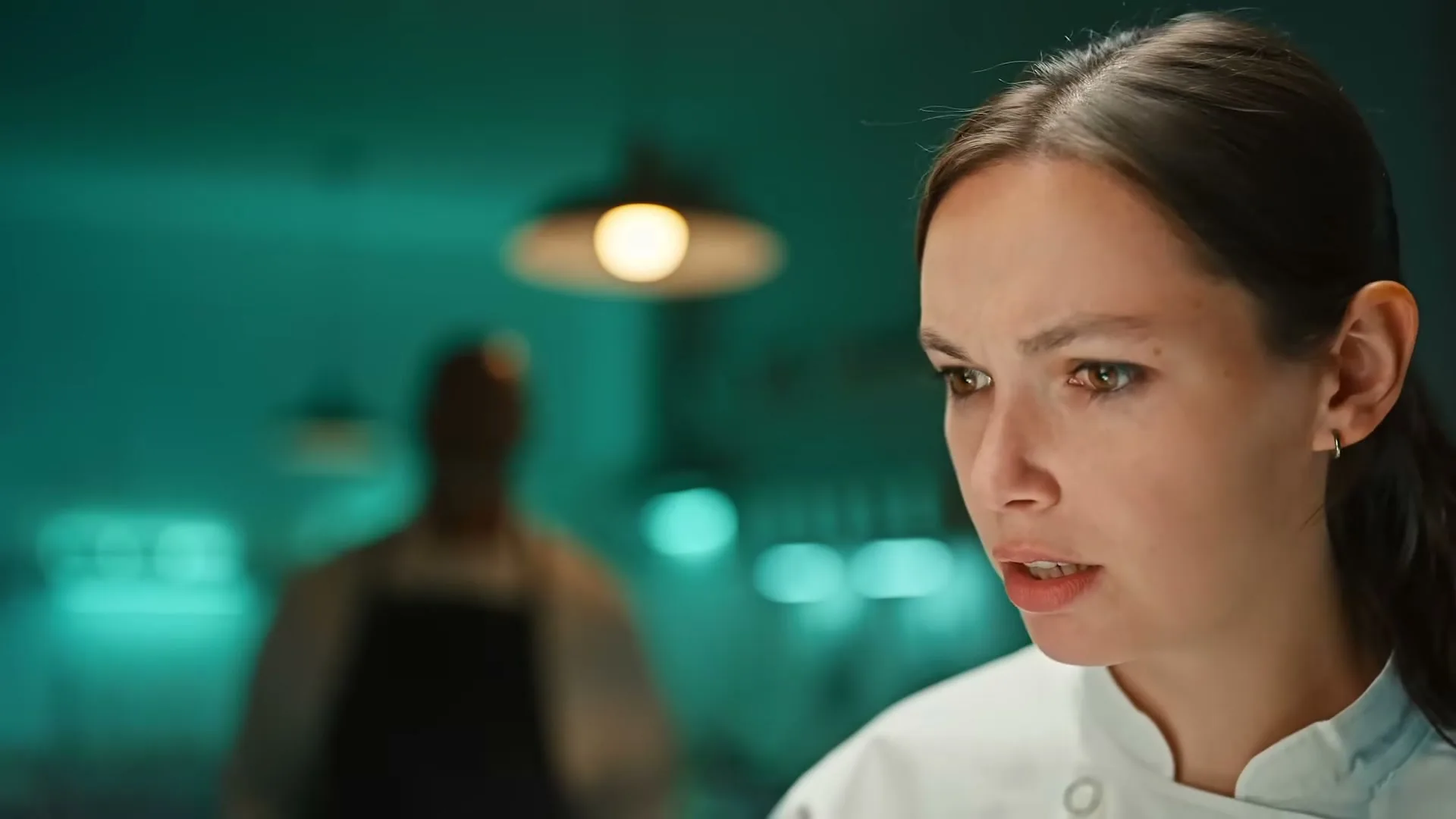 A female chef in a white uniform looks surprised, with a blurred male figure in an apron in the background of a restaurant kitchen.
