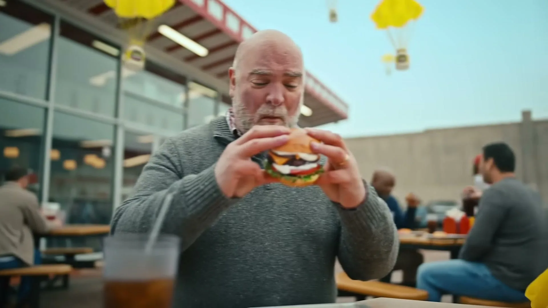 A woman in a purple cardigan holds a jar of mayonnaise and a drink, looking up at parachuting mayonnaise jars, then a man holds a burger, looking at it intently at an outdoor restaurant.