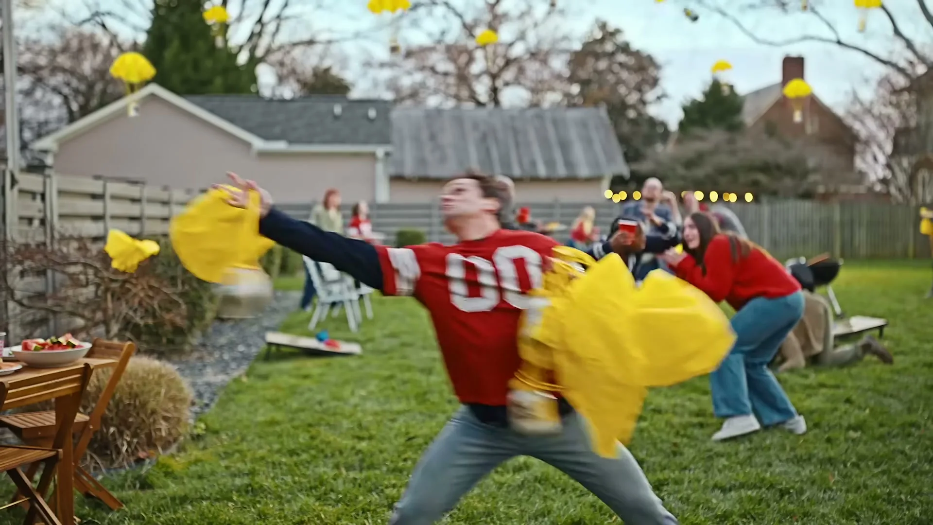 A young man in a red football jersey is hit by yellow parachutes in a backyard full of people playing games.