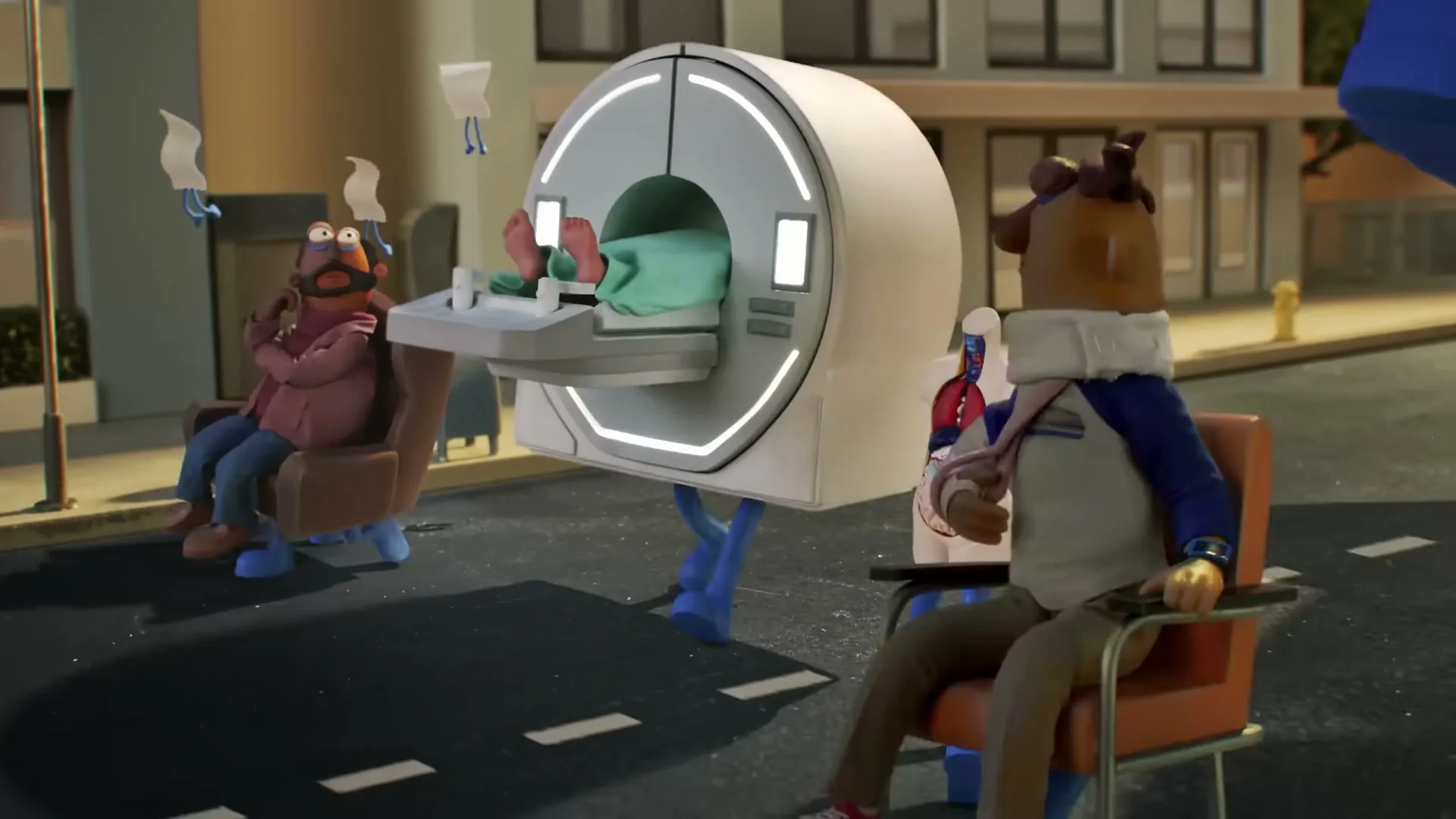 Two claymation men in chairs and an MRI machine with a patient's feet visible, all with legs, move along a street in front of a building.