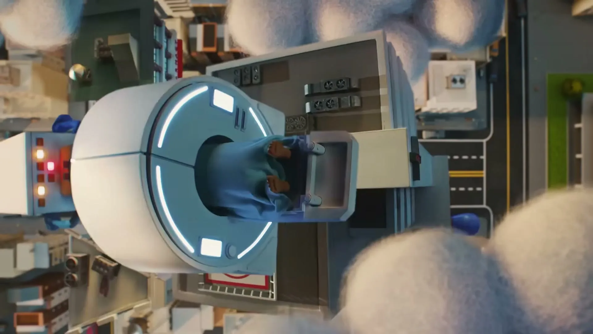 An overhead shot shows a toy-like hospital building with a patient being scanned in an MRI machine, surrounded by fluffy clouds and a miniature city.