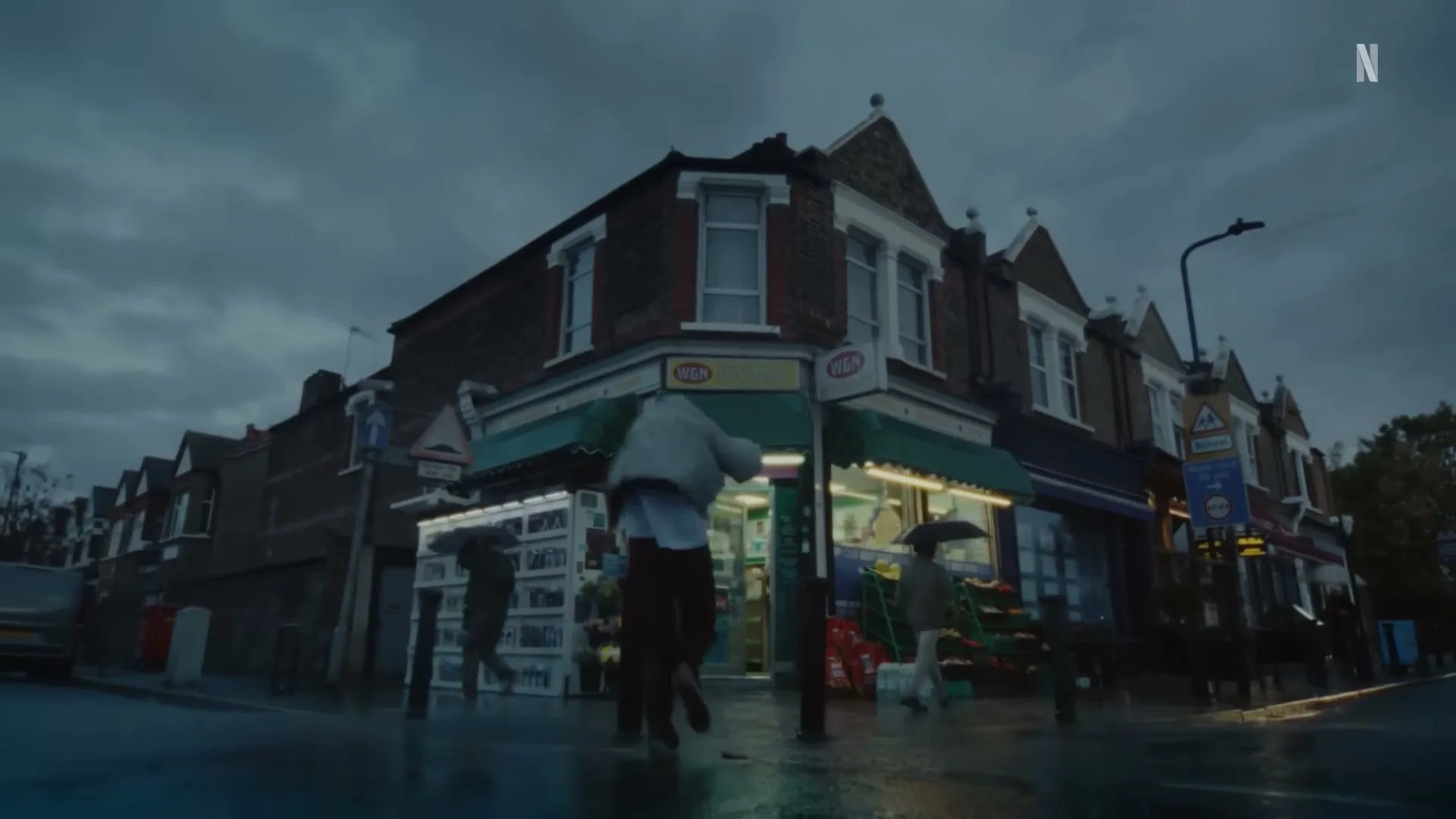 A person in a light jacket and dark pants runs into a brightly lit corner store on a rainy street at dusk, with other people holding umbrellas nearby.