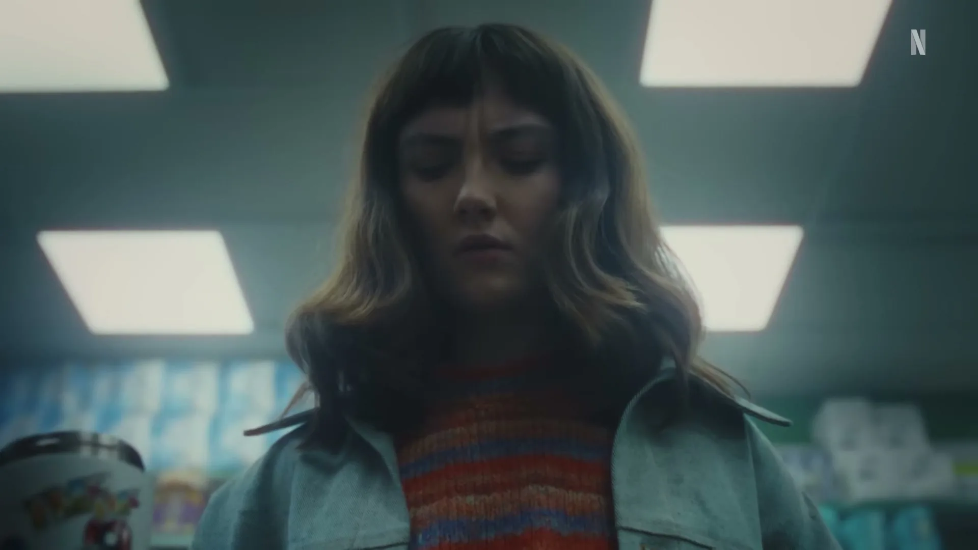 A young woman with brown hair and bangs, wearing a striped sweater and denim jacket, looks down intently while holding a can in a brightly lit store.