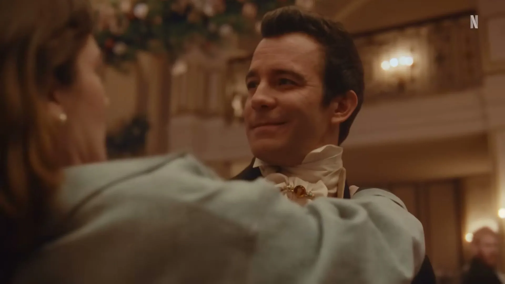 A man in period costume smiles at a woman in a ballroom, as they dance together in a warm, elegant setting.
