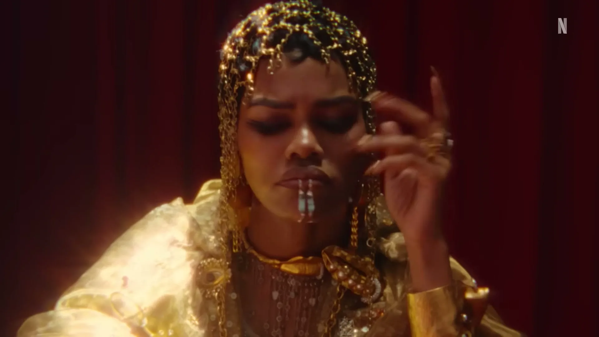 A woman with a golden headpiece and ornate clothing holds two cigarettes in her mouth, looking down thoughtfully in a dimly lit room.